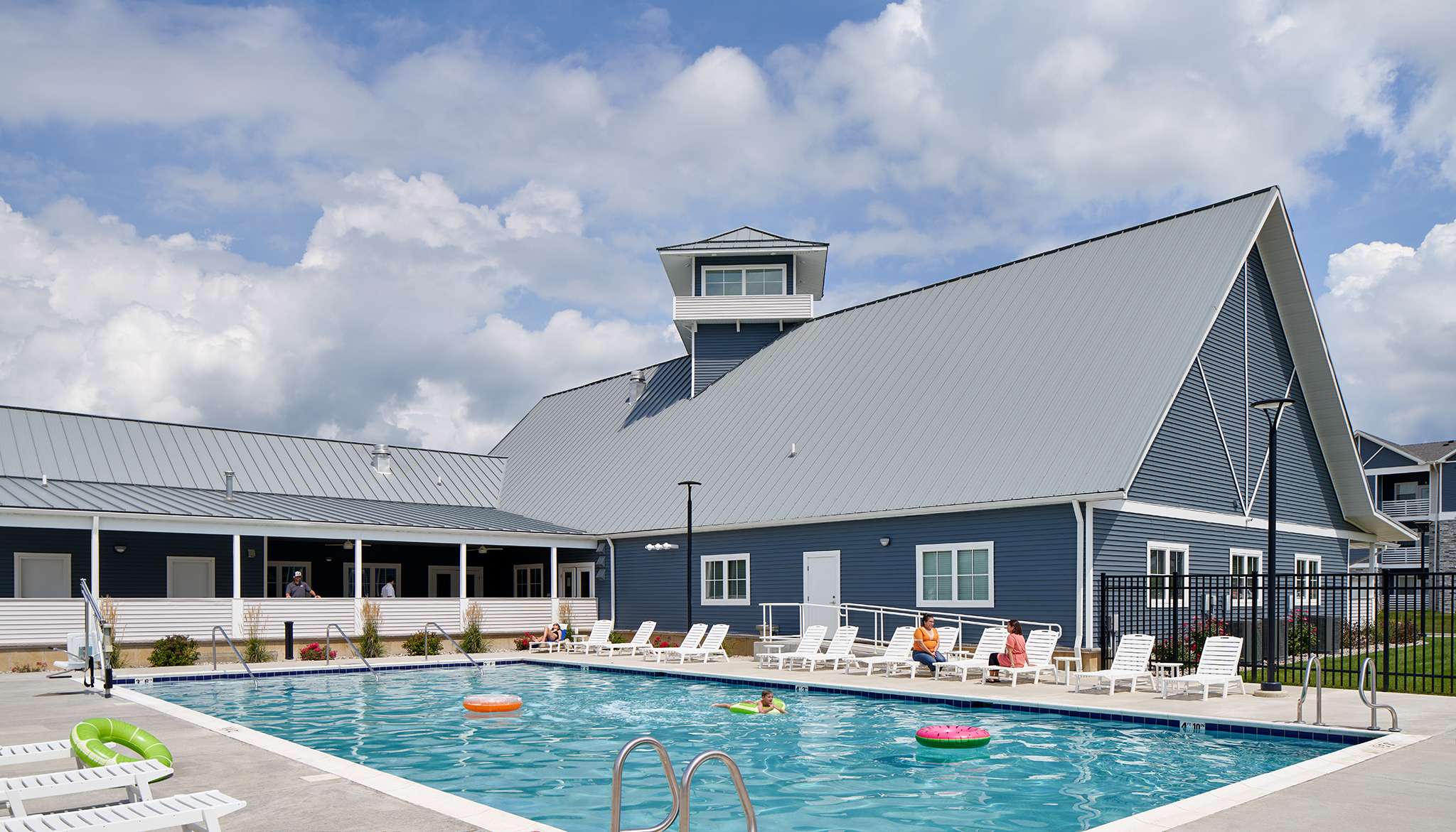 Clubhouse with outdoor swimming pool and a few people swimming/sitting on the deck.